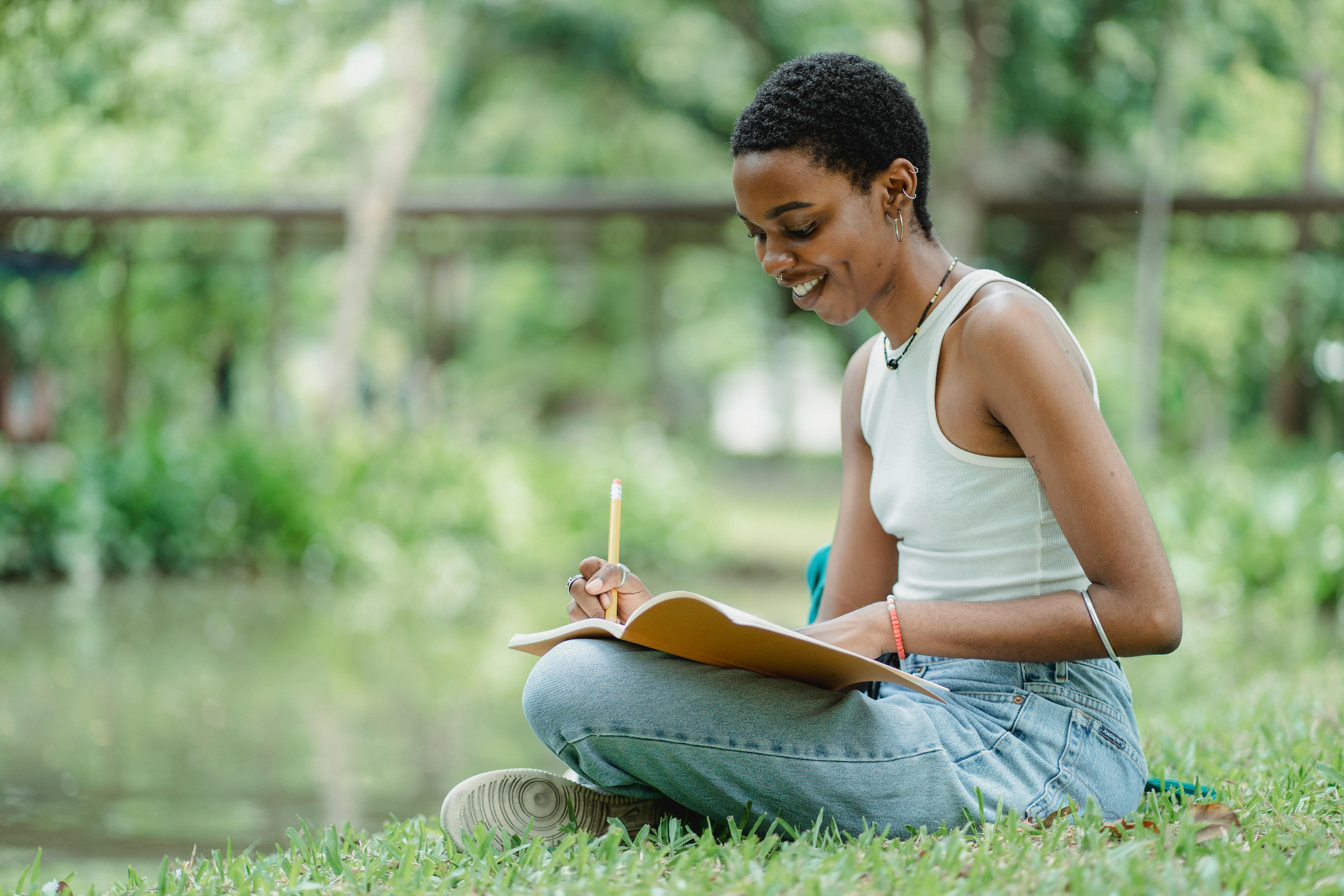 black-female-writing-in-notepad-while-sitting-on-meadow